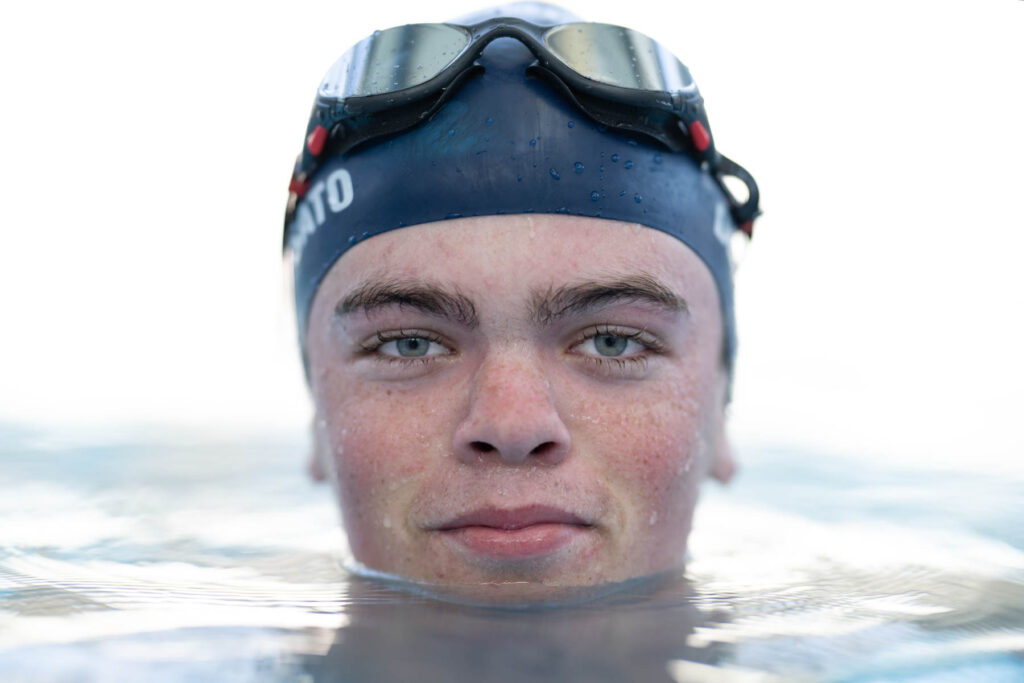 Up-close photo of a high school aged person poking their head out of the water while wearing a swim cap and goggles on top of their head.
