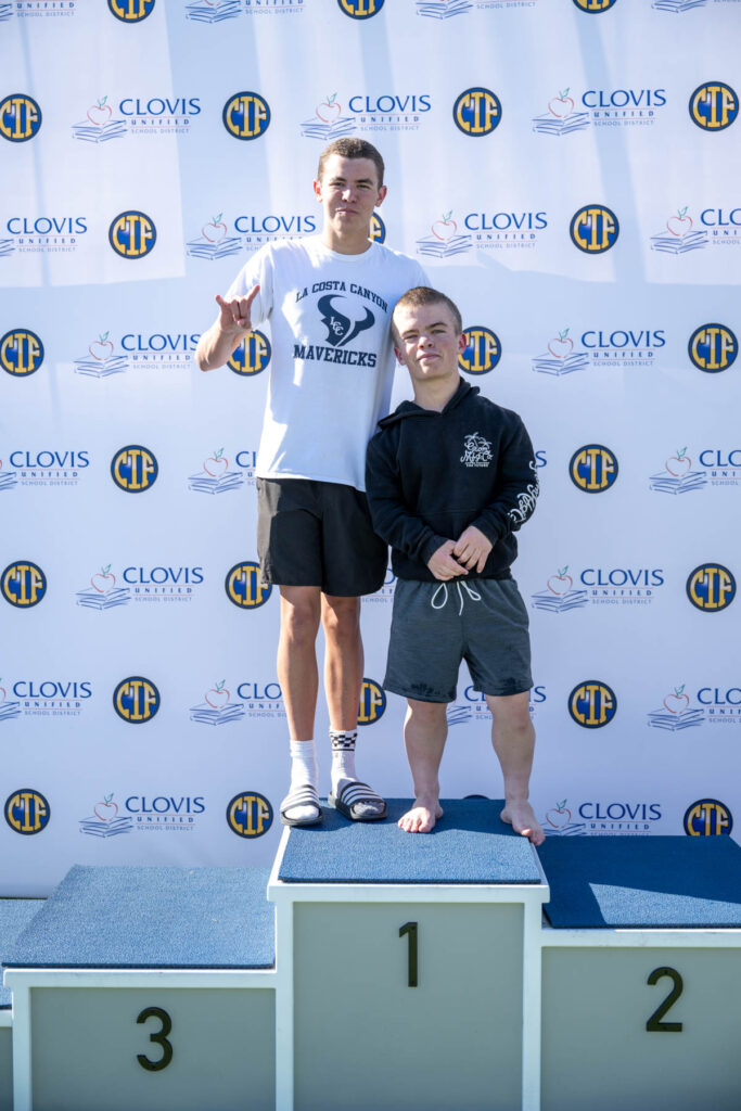 Two people, one of whom has dwarfism, standing on podium 1 against a CIF backdrop and smiling at the camera.