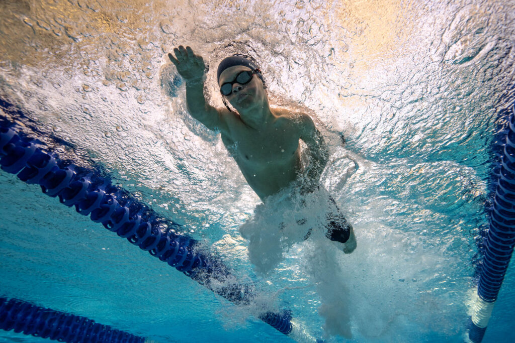 An underwater full-body photo of a person with dwarfism swimming with the surface above them.