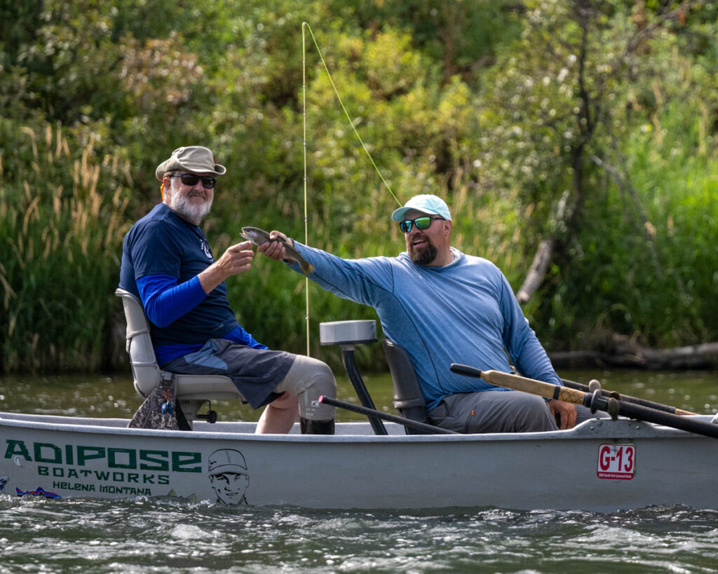 Two people in a boat; one person is holding a fish out to the other person, who is pointing at it and smiling at the camera.
