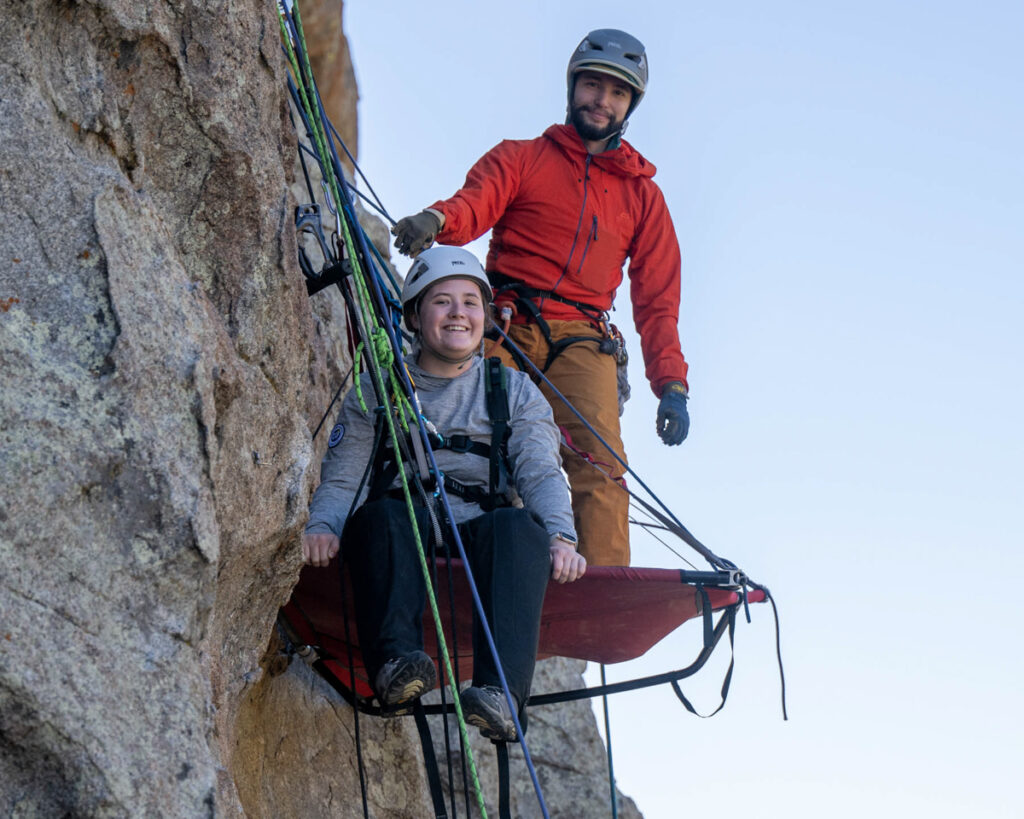 Two climbers secured on a cliff, with one sitting on a red portaledge and the other standing.