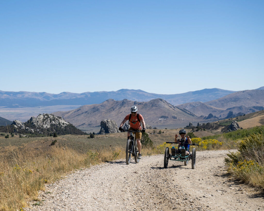 An upright cyclist and a person in a recumbent tricycle riding along a gravel path in a mountainous landscape under a clear blue sky.