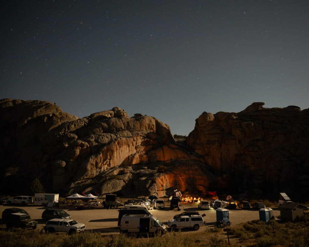 A camping area with vehicles and a campfire near large rocky formations under a starry night sky.