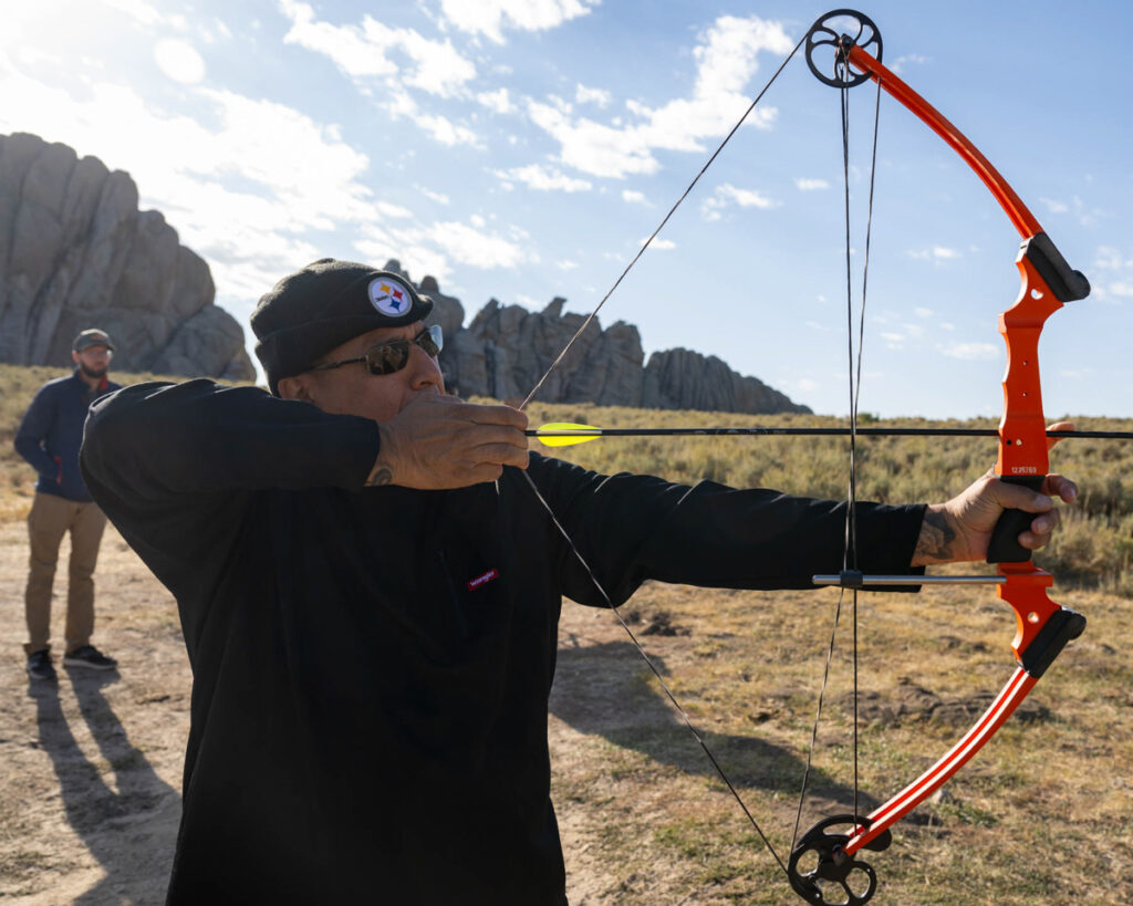 A person aiming an orange bow with a yellow arrow outdoors, with rock formations in the background.