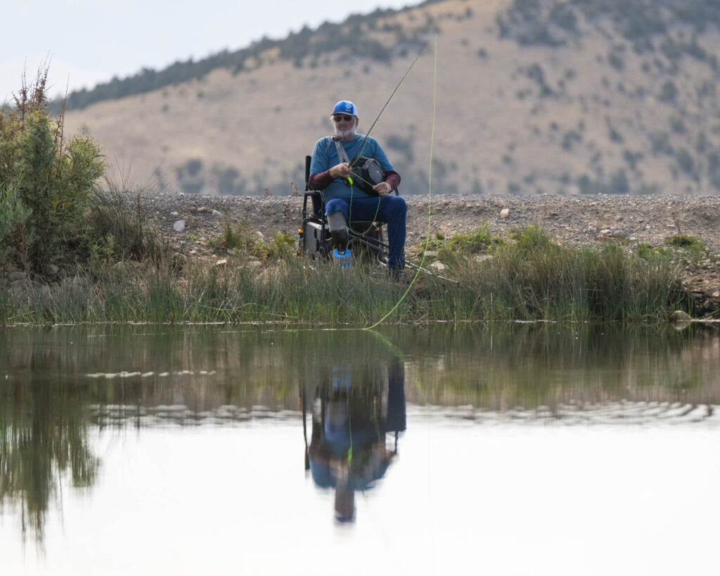 A person in a wheelchair fishing by a tranquil body of water with a hilly background.