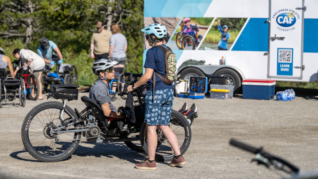 People gathered around adaptive tricycles and a trailer with the "Challenged Athletes Foundation" logo.