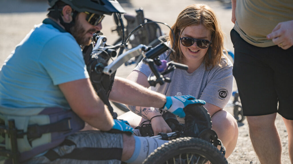 A group of people gathered around an adaptive bicycle outdoors, with one person adjusting the wheel.