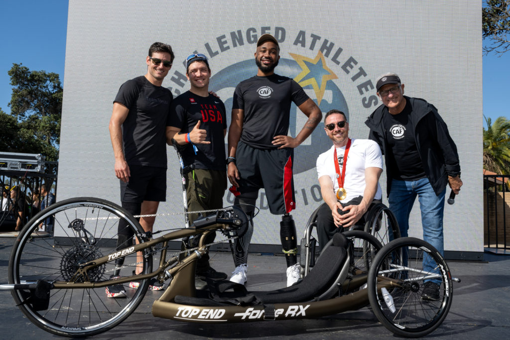 A group of five athletes, including wheelchair and prosthetic users, poses on stage with a racing handcycle in front of the CAF logo.