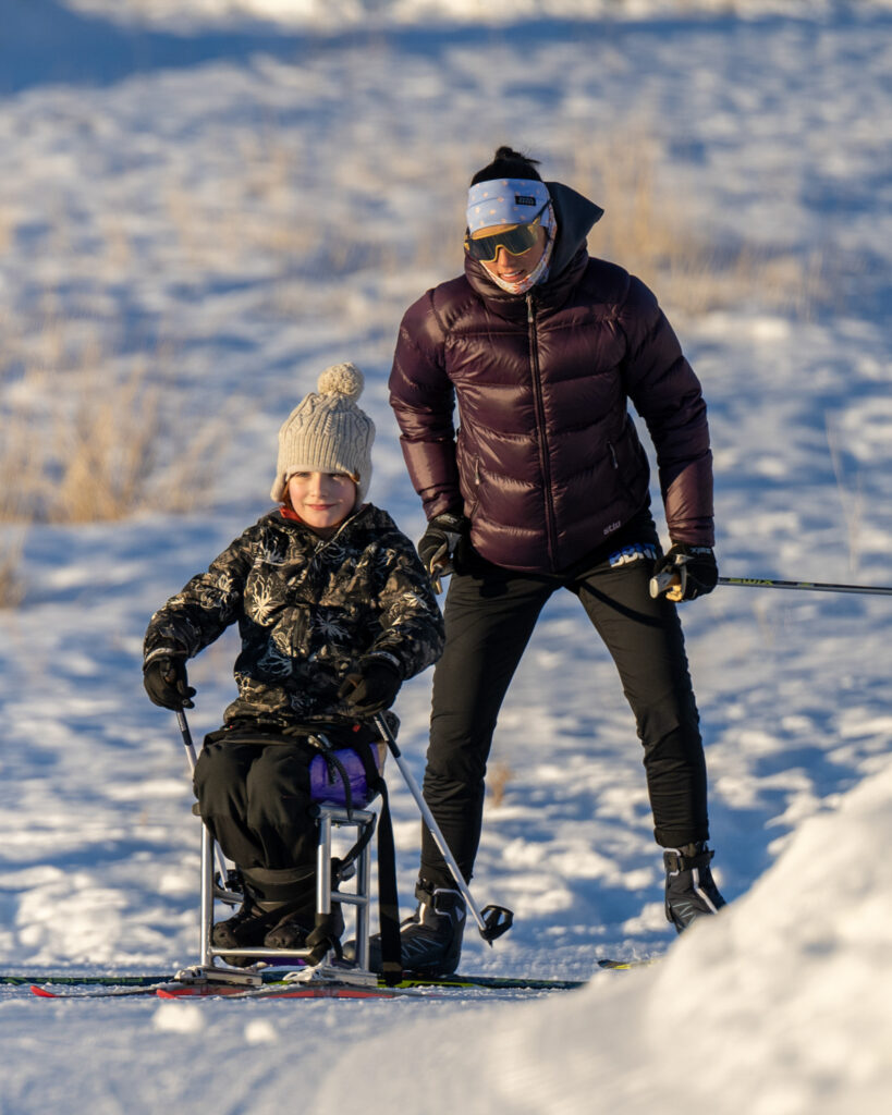 A child in a sled on snow, pushed by an adult in winter clothing.