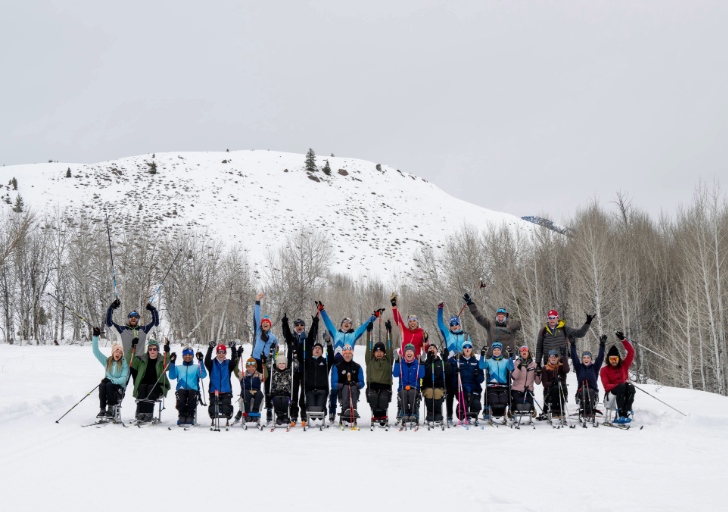 A large group of people wearing ski gear in a snowy meadow next to a snowy mountain. Many are in sit skis and are gesturing excitedly into the air.
