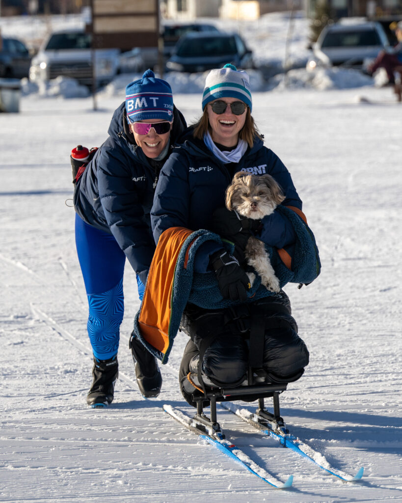Two people enjoying sledding across snow, with one seated in a sit ski and holding a dog, and the other pushing the sit ski.