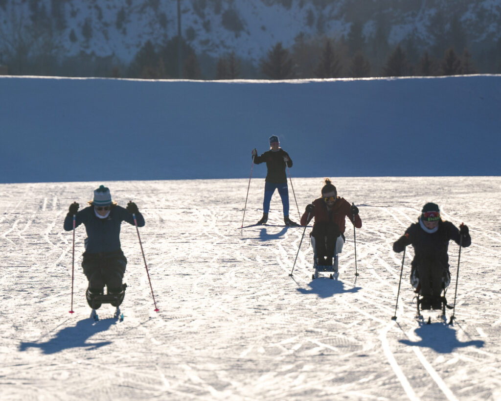 Four people skiing on a snowy landscape, three using sit-skis and one standing with poles.