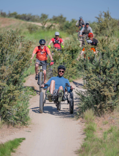 Athlete using an adaptive mountain bike riding a dirt trail alongside cyclists at CAF Idaho Mountain Bike Weekend