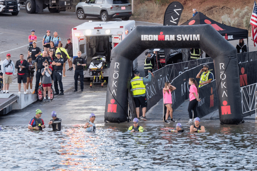 Athletes wearing swim caps wait in the water near the IRONMAN 70.3 swim start arch, surrounded by event staff and spectators.