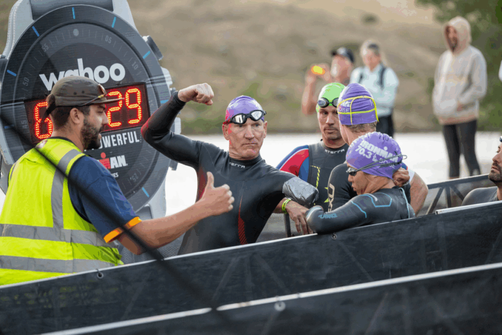 Swimmers in wetsuits and purple caps gather at the start line, one athlete flexing his arm while a race official gives a thumbs up.