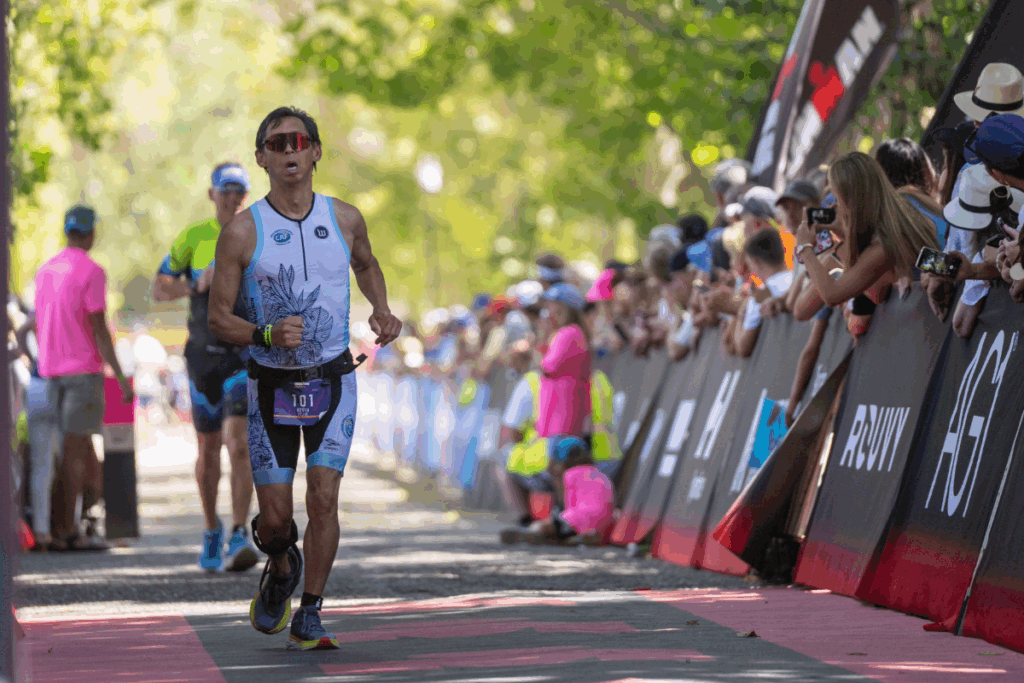 A runner approaches the IRONMAN 70.3 Boise finish line, focused as spectators line the course under shaded trees.