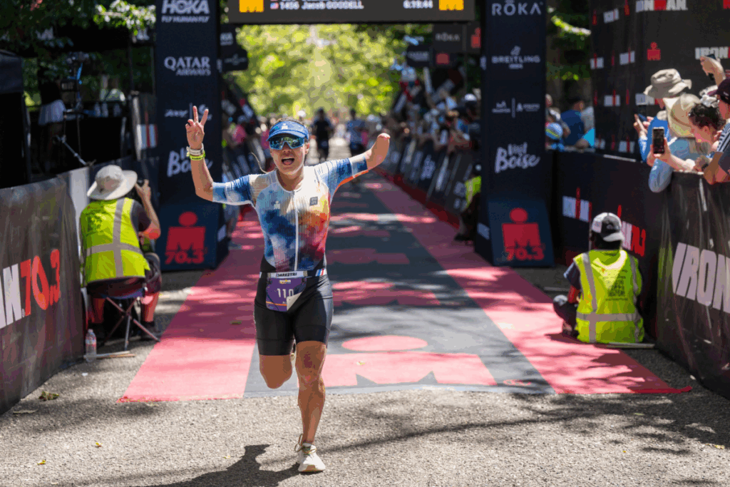 A smiling runner raises both arms in celebration as they cross the IRONMAN 70.3 Boise finish line, surrounded by cheering spectators and race staff.