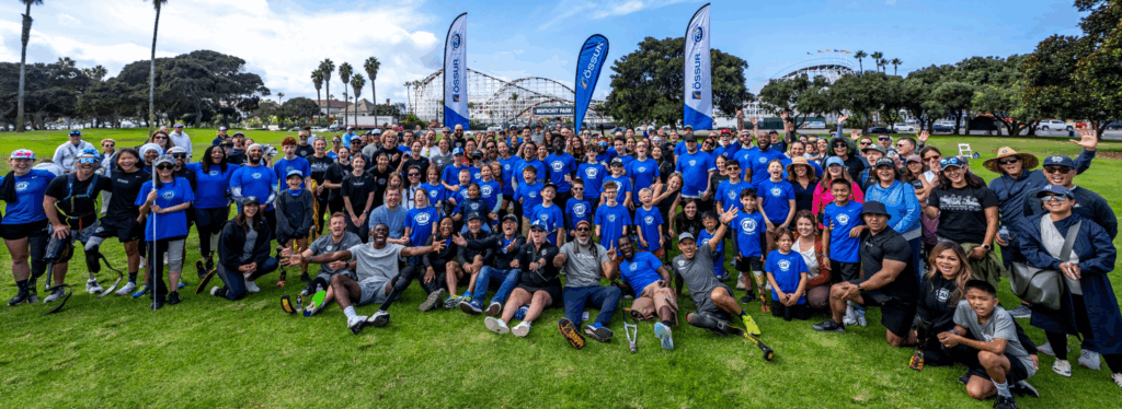 Large group of athletes, volunteers, and families in CAF shirts gathered on the grass with roller coasters in the background at San Diego Triathlon Challenge weekend.