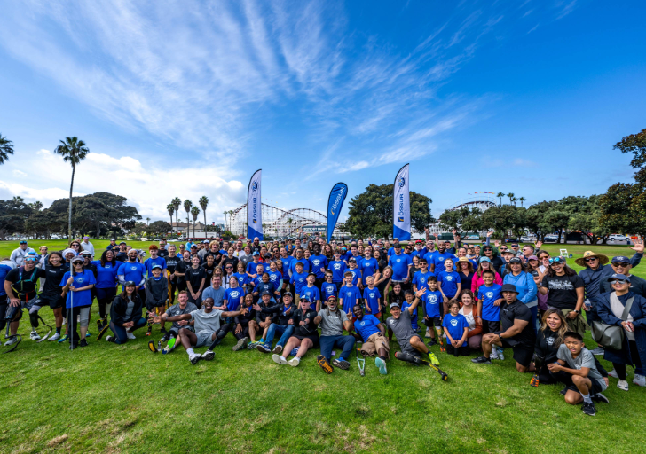Large group of athletes, volunteers, and families in CAF shirts gathered on the grass with roller coasters in the background at San Diego Triathlon Challenge weekend.