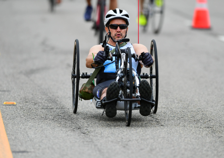 An athlete with a physical disability powers a handcycle along the course during the Zuma Beach Triathlon, wearing a helmet and sunglasses while competing on a closed roadway.