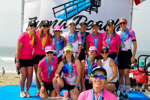 A team of women wearing medals and bright pink tops pose together on the beach at the Zuma Beach Triathlon, celebrating their race accomplishment with the ocean behind them.
