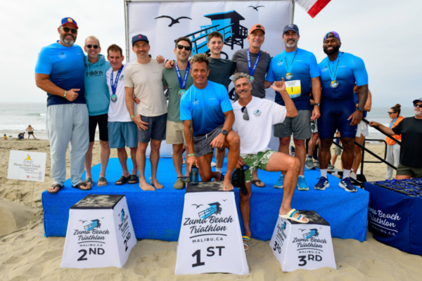 Athletes and supporters stand on the awards podium at the Zuma Beach Triathlon, smiling and holding medals after the race in Malibu.