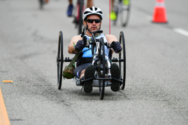 An athlete with a physical disability powers a handcycle along the course during the Zuma Beach Triathlon, wearing a helmet and sunglasses while competing on a closed roadway.