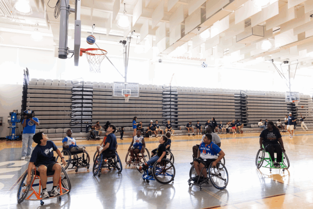Youth athletes using sport wheelchairs aim for the basketball hoop during a game in a gym, with spectators and teammates watching.
