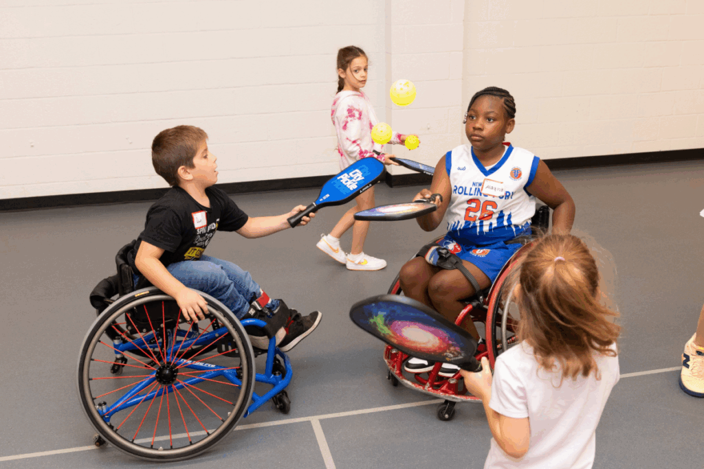 oung athletes, including two wheelchair users, play a game of pickleball indoors, practicing balance and coordination with paddles and balls.