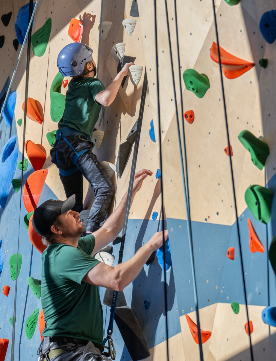 Young athlete with a limb difference climbs an indoor rock wall with the support of a coach providing a belay
