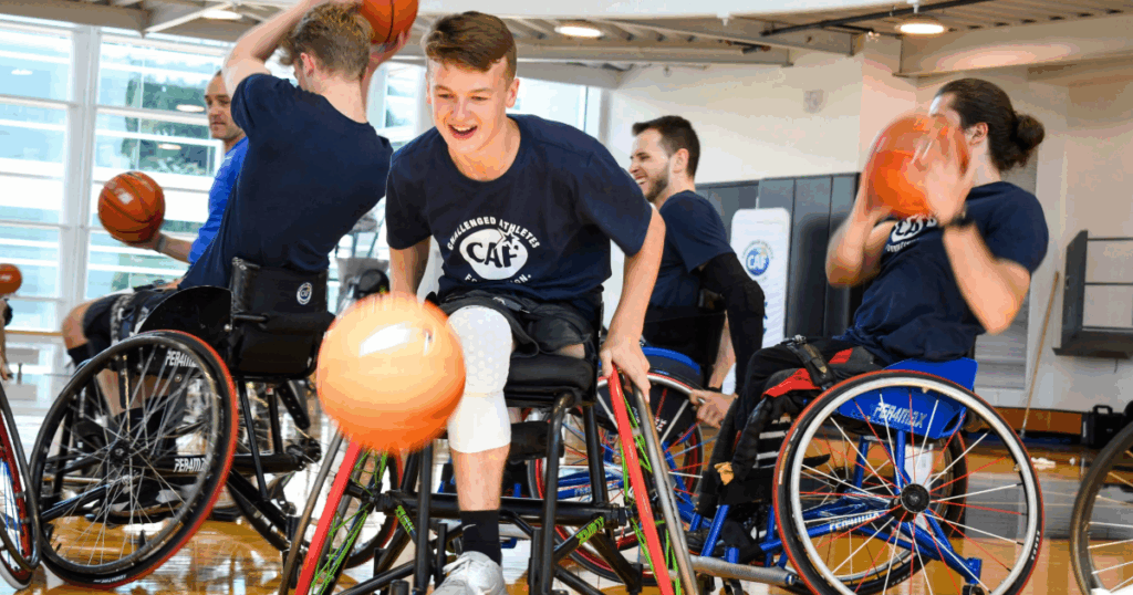 Athletes in sport wheelchairs play basketball indoors, smiling and engaged in the game during a clinic