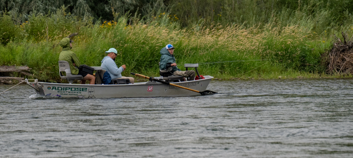 Three anglers sit in a drift boat fly fishing on a calm river surrounded by tall grass and trees.