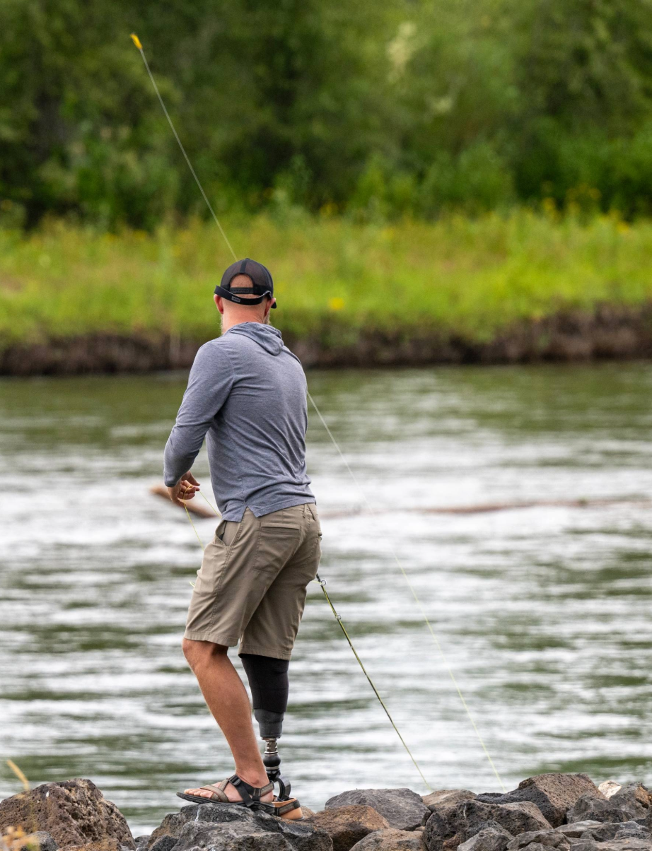 An angler with a prosthetic leg casts a fishing line while standing on rocks along a riverbank.