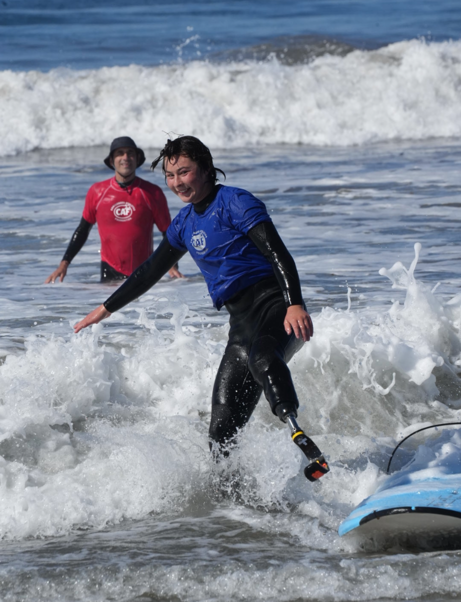 Athlete with a prosthetic leg surfing with a smile as a CAF volunteer supports from behind.