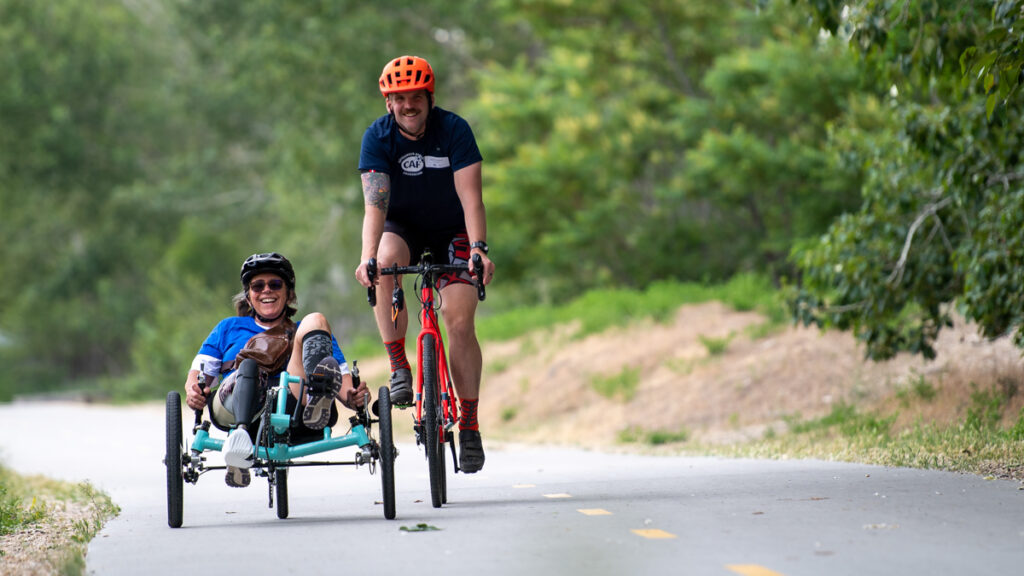 Two people riding together on a path, one on a recumbent tricycle and the other on a bicycle, surrounded by greenery.
