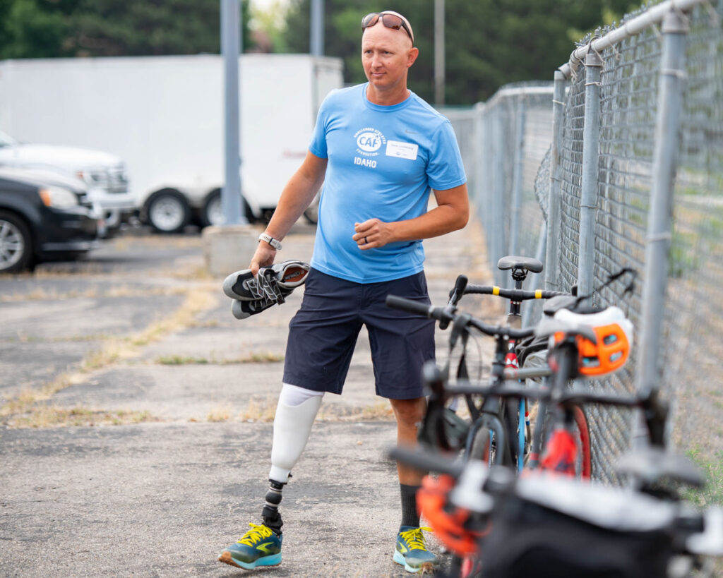 A person with a prosthetic leg walking near parked bicycles and a fence, holding shoes and wearing a light blue T-shirt.