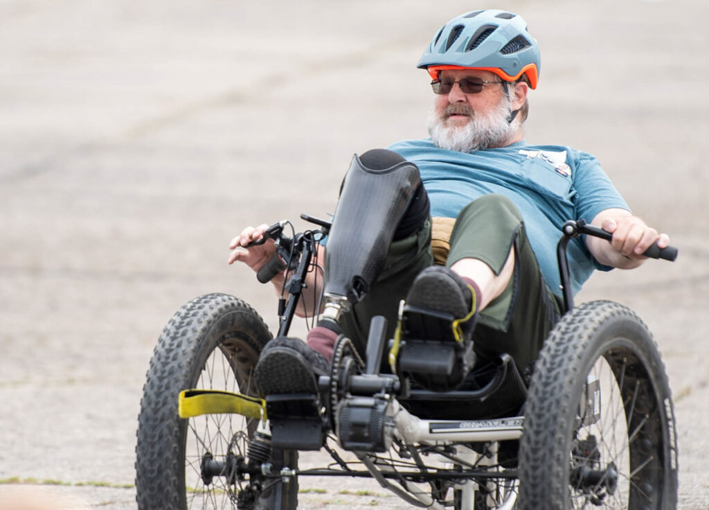 Man with a beard riding a recumbent tricycle on a paved surface, wearing a blue helmet and shirt, with a prosthetic leg.