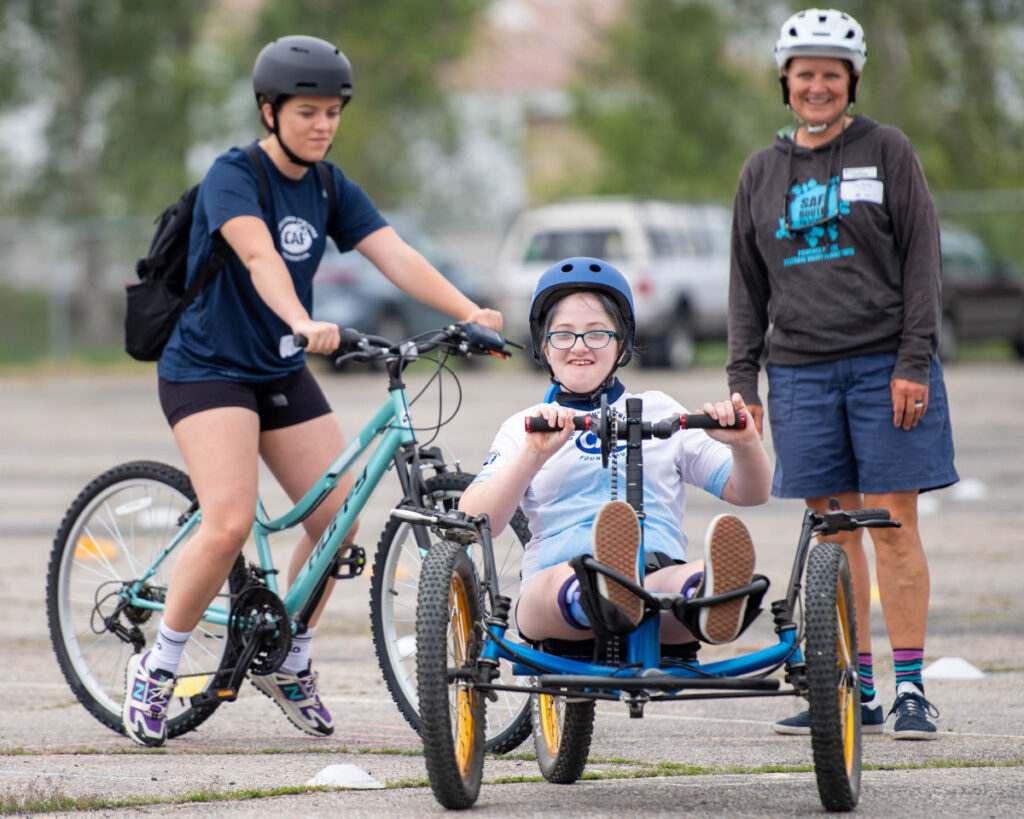Three people at a cycling event, with one on a recumbent tricycle and two others standing nearby with bikes and wearing helmets.