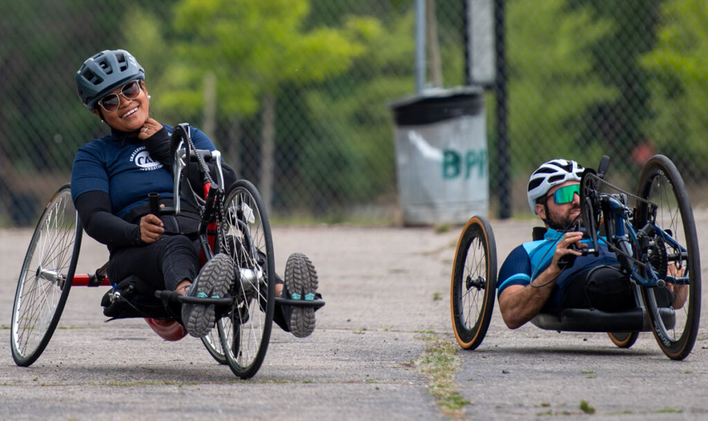 Two athletes on handcycles, wearing helmets and riding on a paved path.