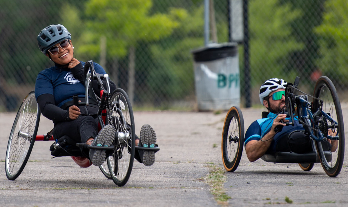 Two athletes on handcycles, wearing helmets and riding on a paved path.