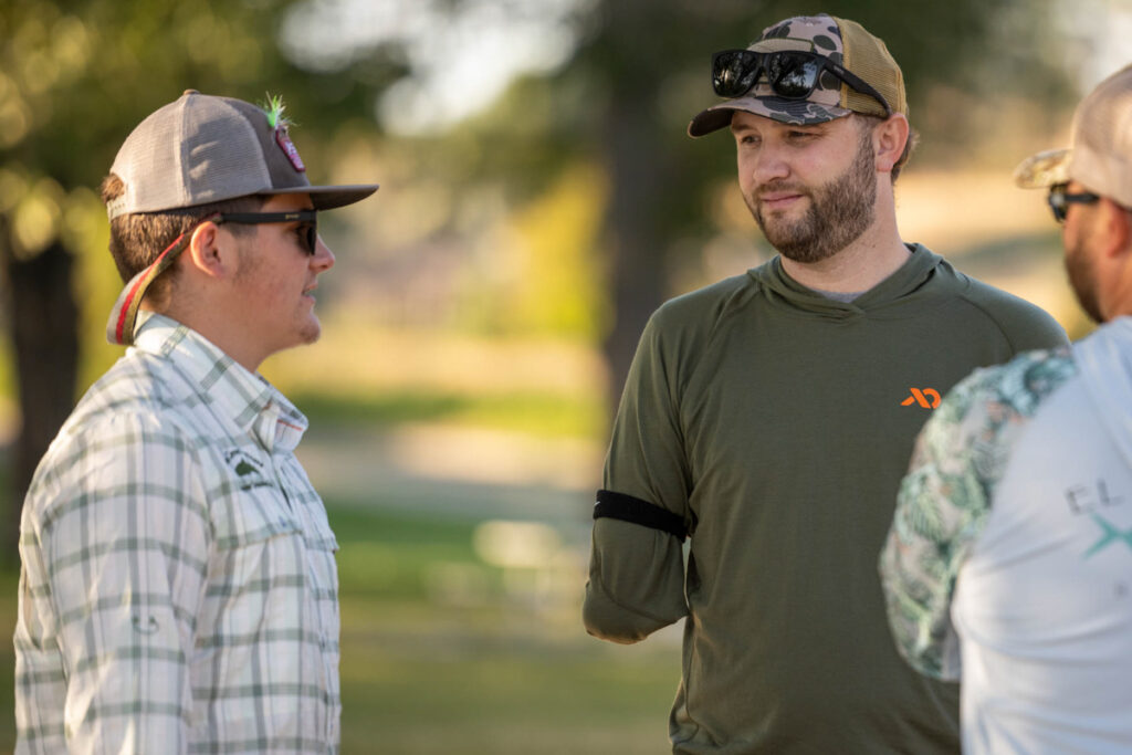 Two men conversing outdoors; one has a plaid shirt and fly-fishing cap, the other wears an olive shirt and has an upper limb difference.