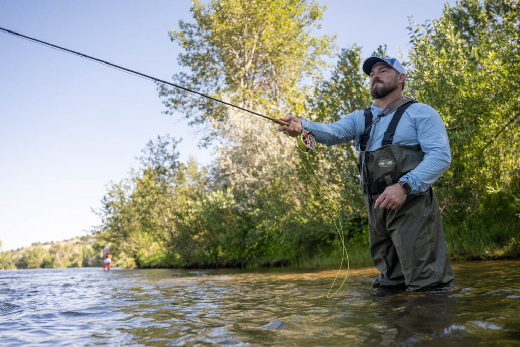 A man in waders fly fishing in a river surrounded by trees.