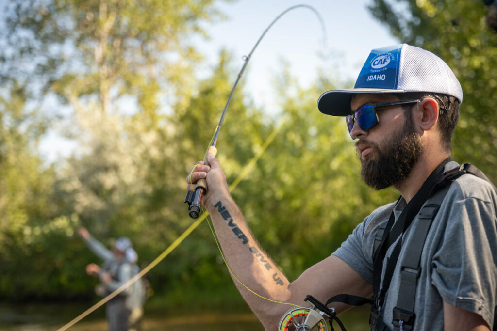 A man fly fishing with a tattoo reading "NEVER GIVE UP" visible on his arm.