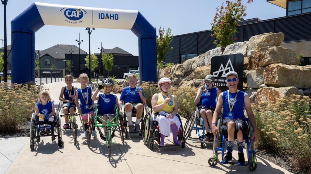 Group of young athletes in wheelchairs display medals under the CAF Idaho finish arch at The Idaho Outdoor Fieldhouse.