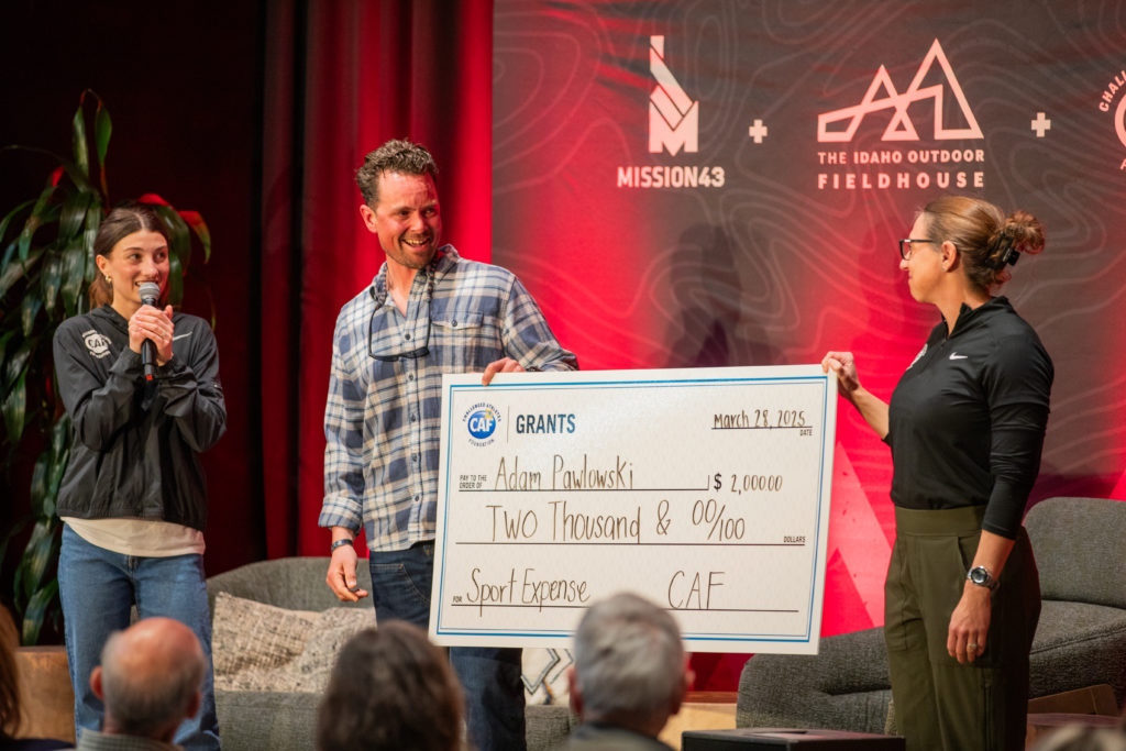 Three presenters on stage hold a large CAF grant check awarded to Adam Pawlowski in Idaho.