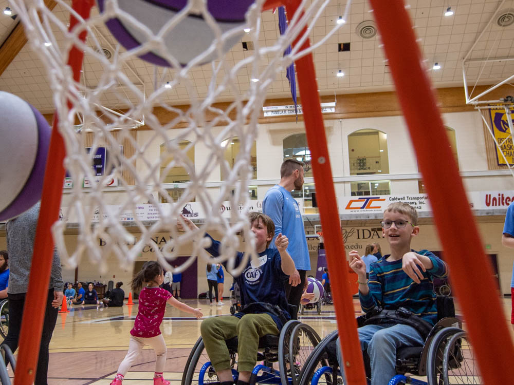 Children in wheelchairs aiming basketballs at a hoop in a gym.