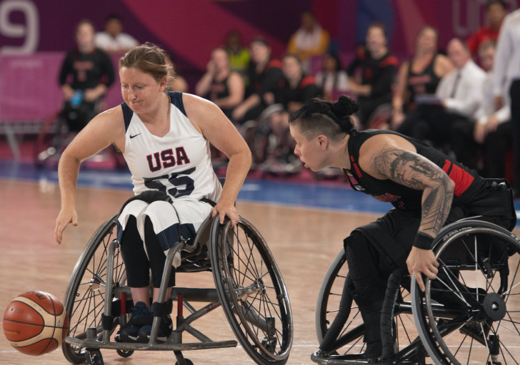Wheelchair basketball players compete for possession of the ball during an international game, with one athlete in a USA jersey and another defender reaching from a sport wheelchair on the court.