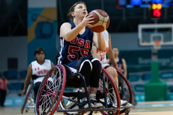 USA wheelchair basketball athlete prepares to shoot a basketball from a sport wheelchair during international competition, with opposing players and a basketball hoop visible in the background.