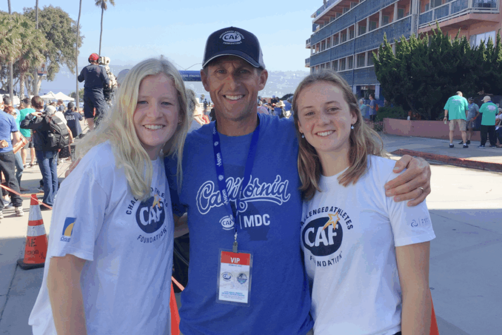 Three people wearing CAF shirts smiling at the San Diego Triathlon Challenge finish area, with spectators and palm trees in the background.