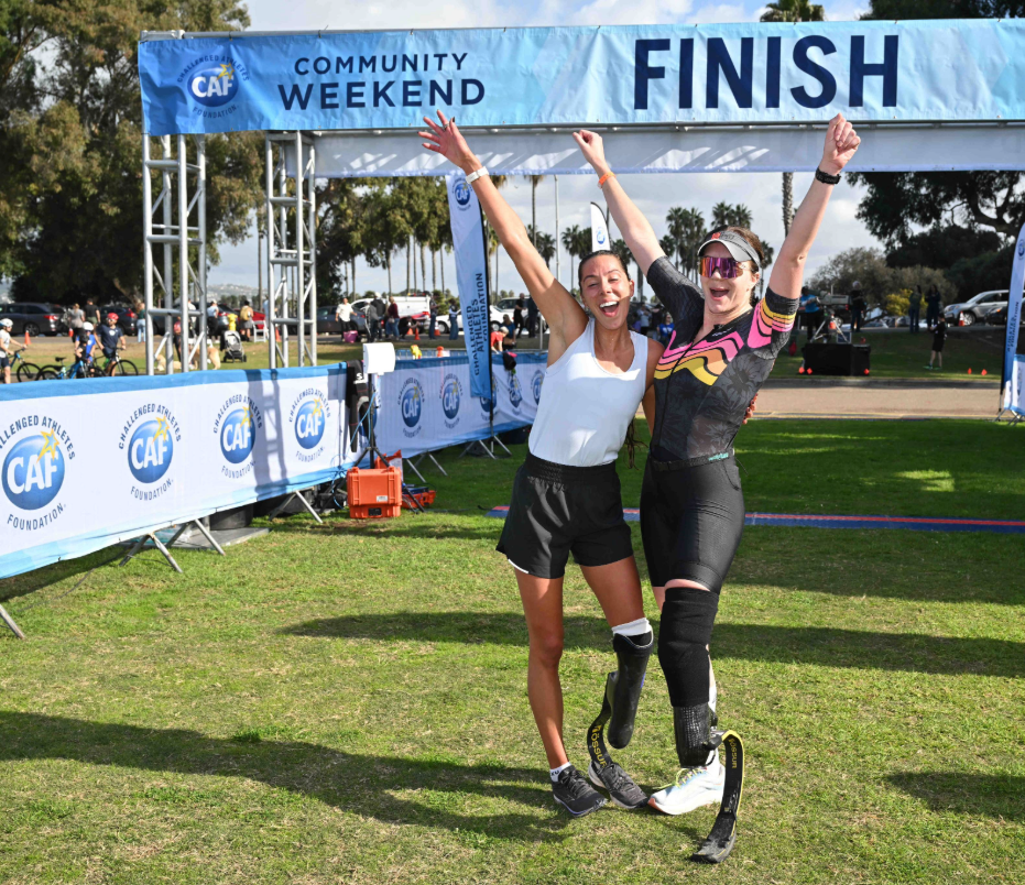 Two athletes with lower limb sports prostheses at the SDTC finish line with hands up in celebratory pose.